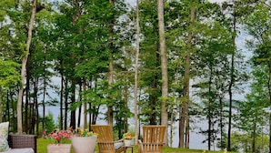 Um terraço com móveis de vime e plantas em vasos, com vista para uma área arborizada e um lago.