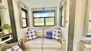 Interior acolhedor de uma motorhome com um sofá de couro bege, duas almofadas xadrez, uma pequena planta em vaso e uma janela com vista para árvores.