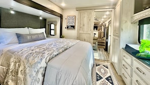 Quarto com uma cama grande, cabeceira, janela e vista para a área da cozinha.