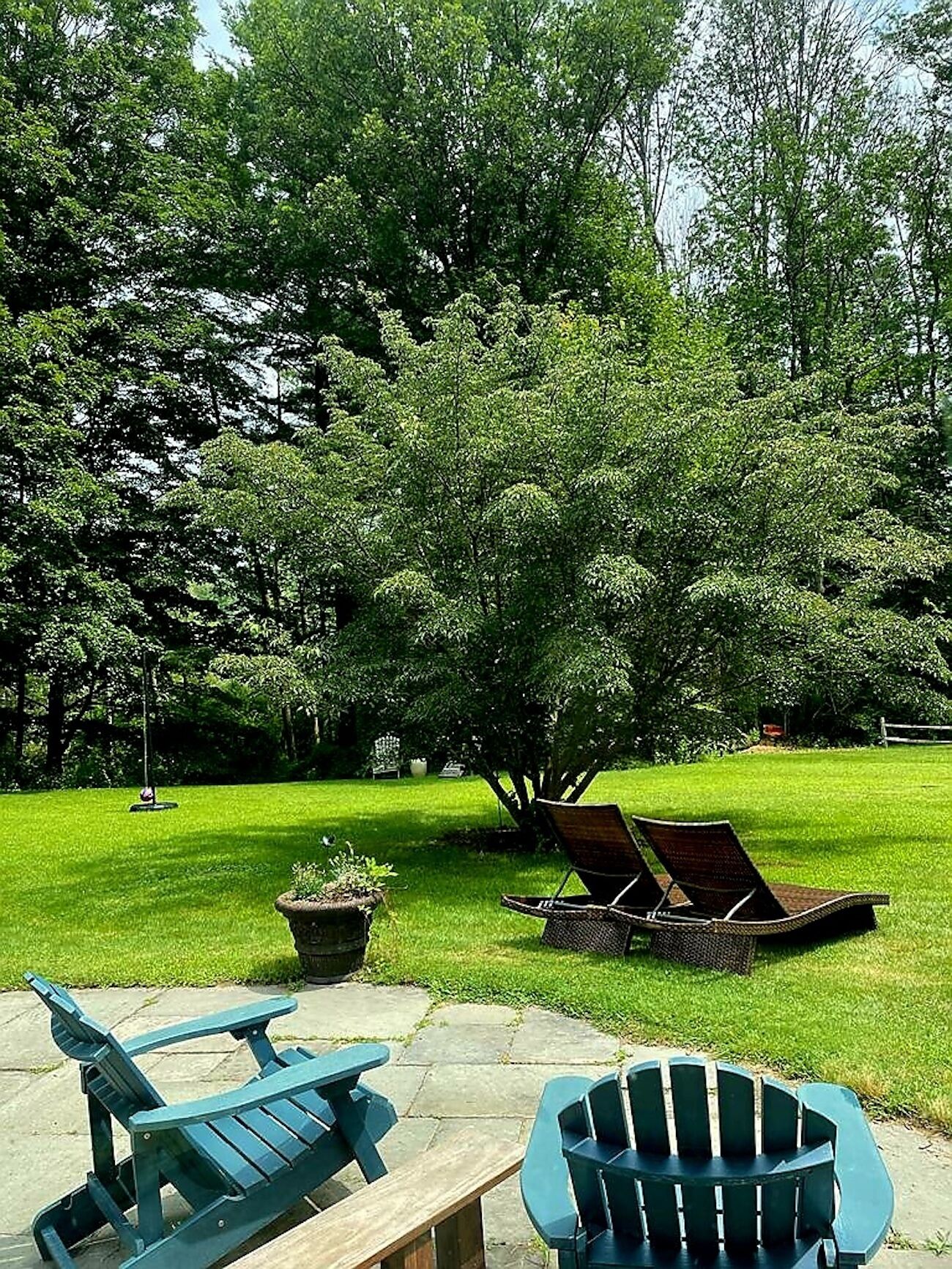 Een terras met Adirondack stoelen en een tafel, omgeven door weelderige begroeiing en bomen.