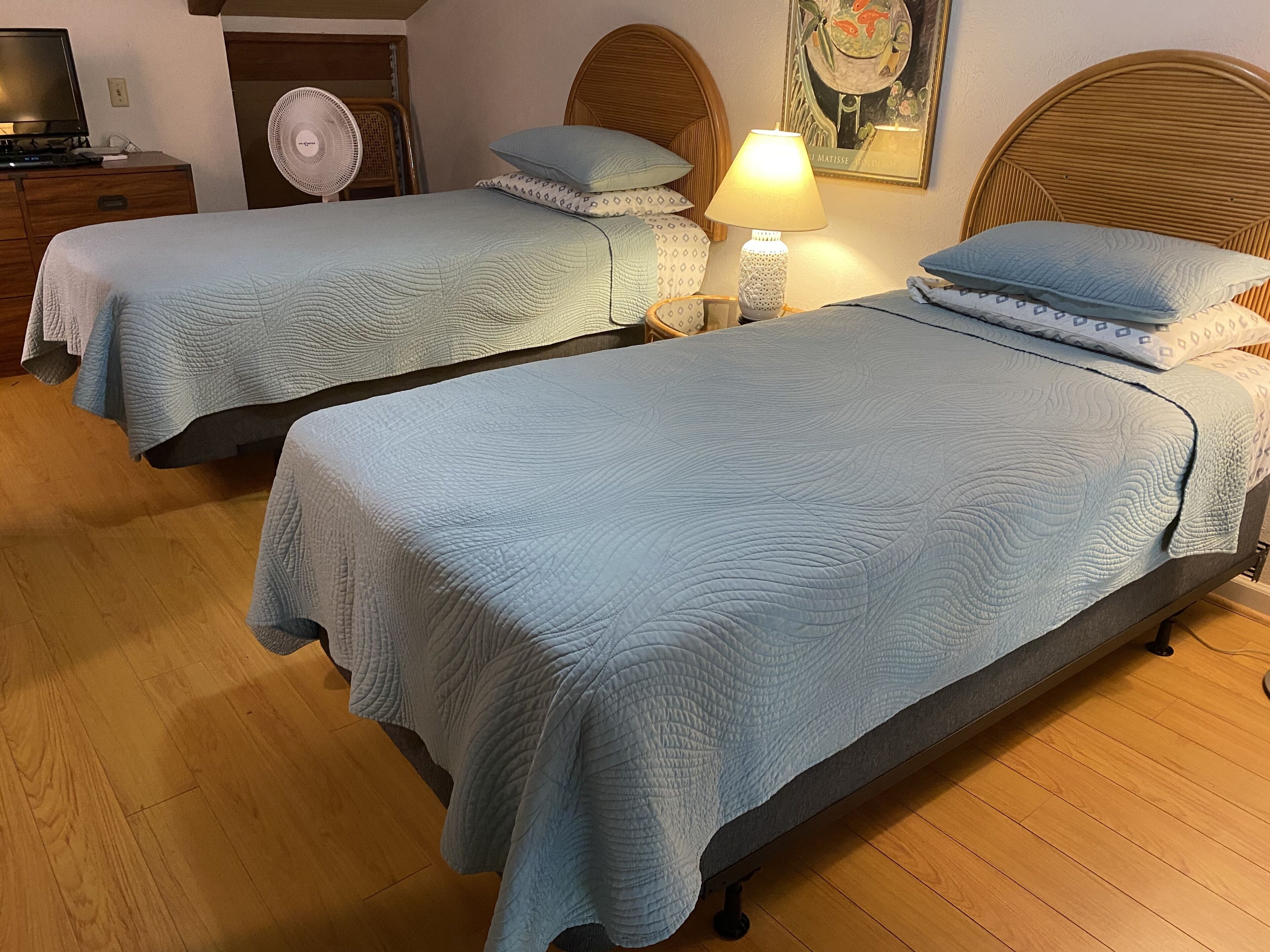 Dos camas individuales con cabeceras y pies de cama a juego, una mesita de noche con lámpara y un ventilador fijado a la pared.