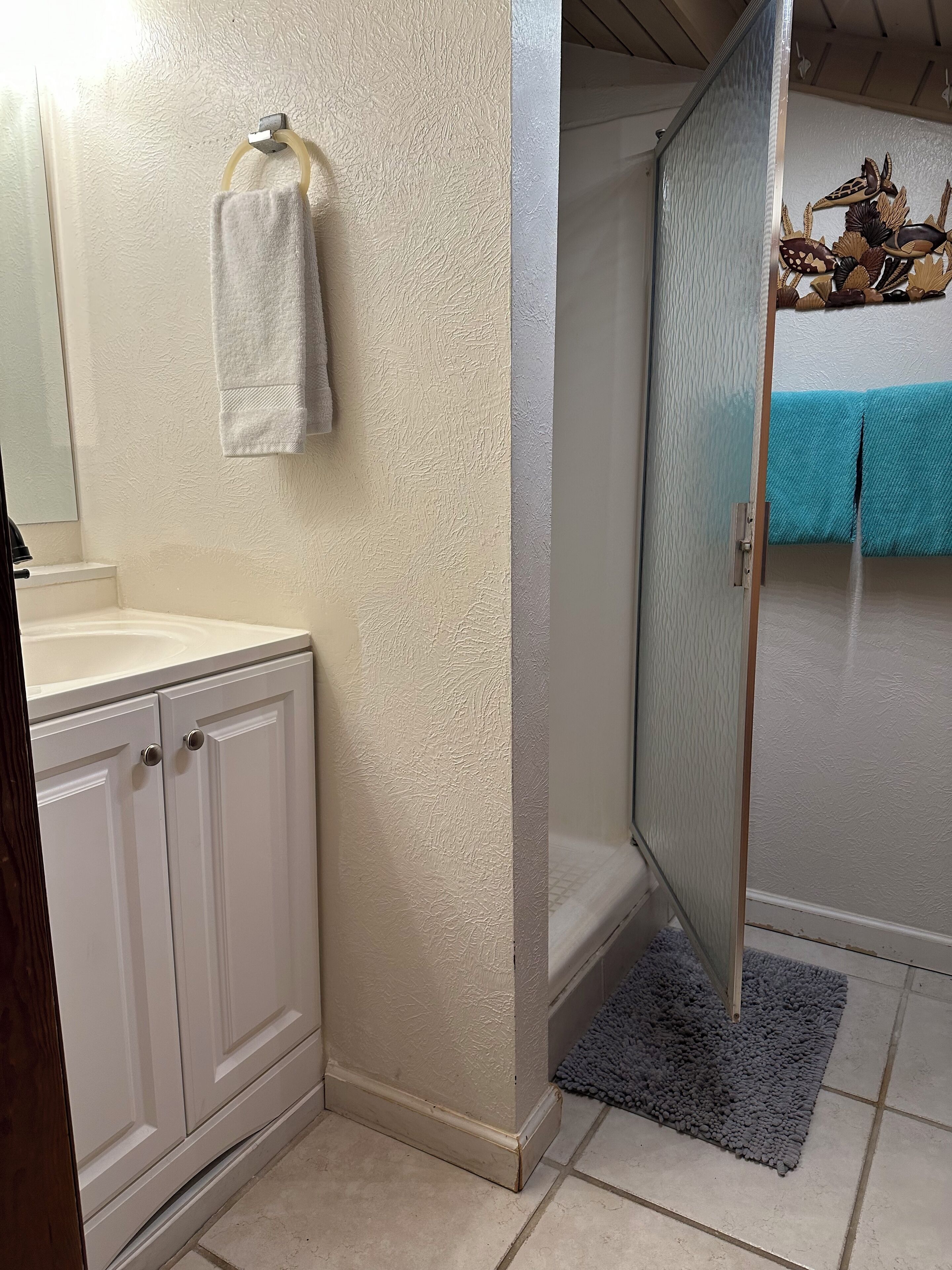 A bathroom with a white textured wall, a white sink, a mirror, a towel rack with a white towel, a shower area with a glass door, and a blue towel on the floor.
