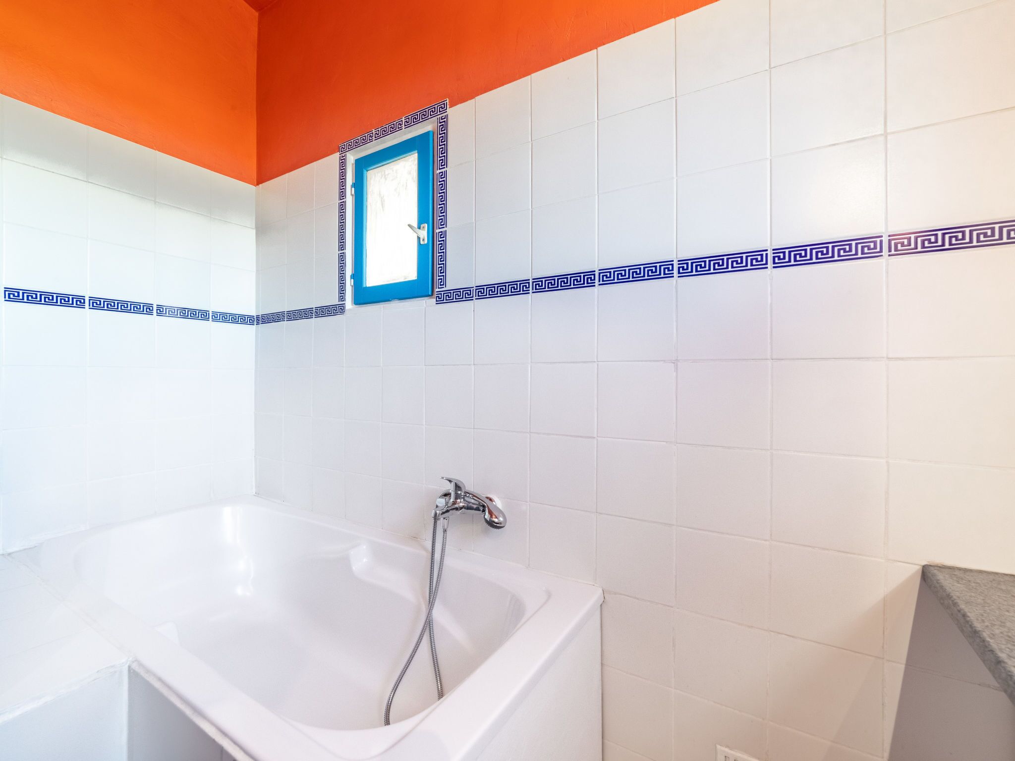 Une salle de bain avec une baignoire, des murs recouverts de carreaux blancs et une fenêtre à cadre bleu.
