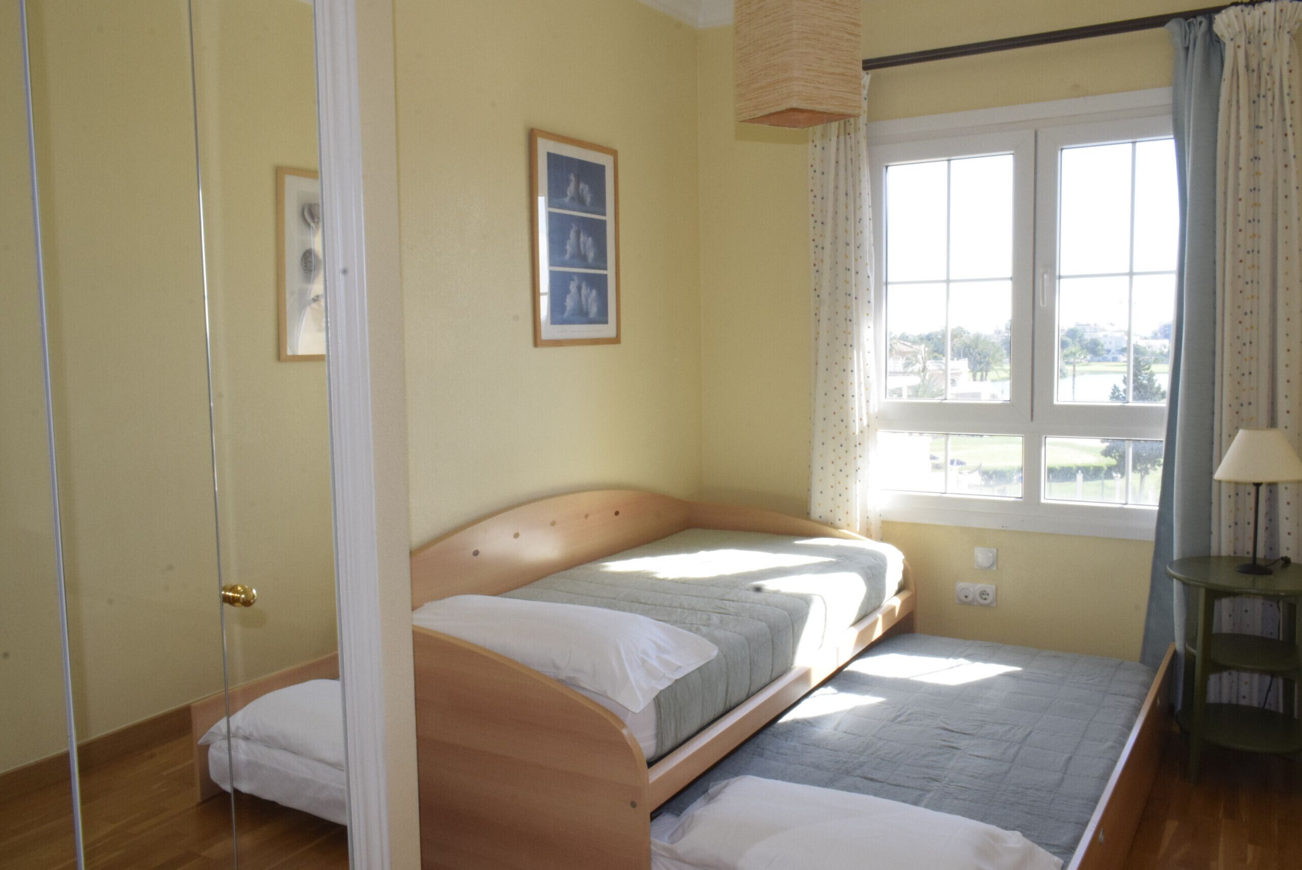 A bedroom with a single bed, a window with curtains, a small table with a lamp, and a framed picture on the wall.