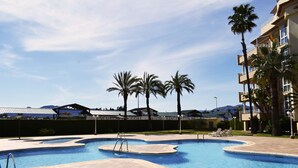 Una piscina con bordeado de baldosas, palmeras y un edificio con balcones al fondo.
