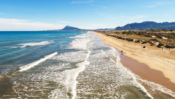 Una playa con olas, una orilla arenosa y montañas a lo lejos.