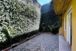 A gravel pathway leading to a yellow building with a door and window, surrounded by dense foliage. - Our Home San Antonio (Cali)
