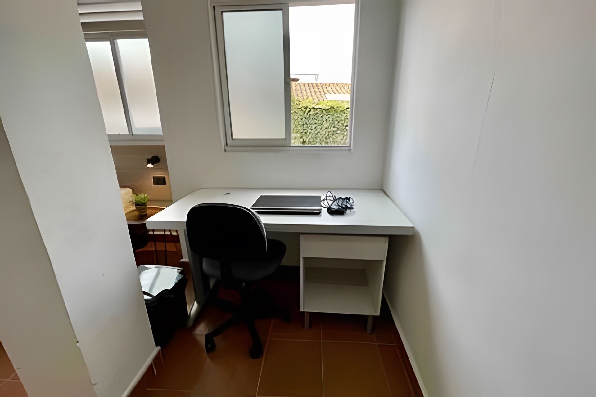 Une petite pièce blanche, avec un bureau, une chaise et une fenêtre.