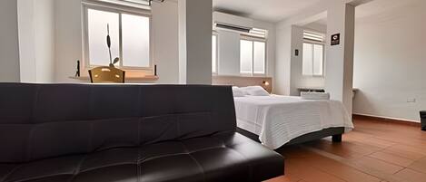 A hotel room with a black sofa, a bed with white bedding, a small table with a lamp, and a window with blinds.