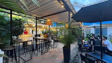 An outdoor cafe area with tables and chairs, surrounded by potted plants and a covered seating area.