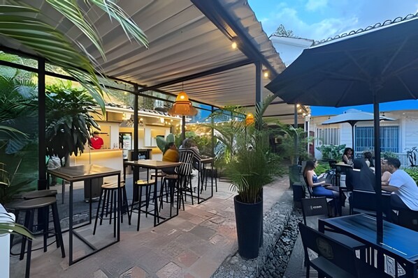 Un área de café al aire libre con mesas y sillas, rodeada de plantas en macetas y una zona de estar cubierta.