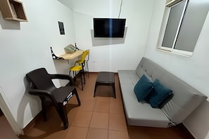 A compact living space with a sofa, a small table, two chairs, a television mounted on the wall, and a window. - Our Home San Antonio (Cali)