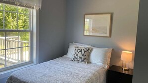 A bedroom with a bed, a pillow with text, a framed picture, a nightstand with a lamp, and a window with a view of greenery. - Abenaki Retreat on Matinicus Island (Matinicus)