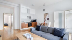 Un apartamento moderno con un sofá gris, suelo de madera, una cocina con armarios blancos y de madera, una zona de comedor con una mesa pequeña y sillas, y un libro sobre la mesa de centro.