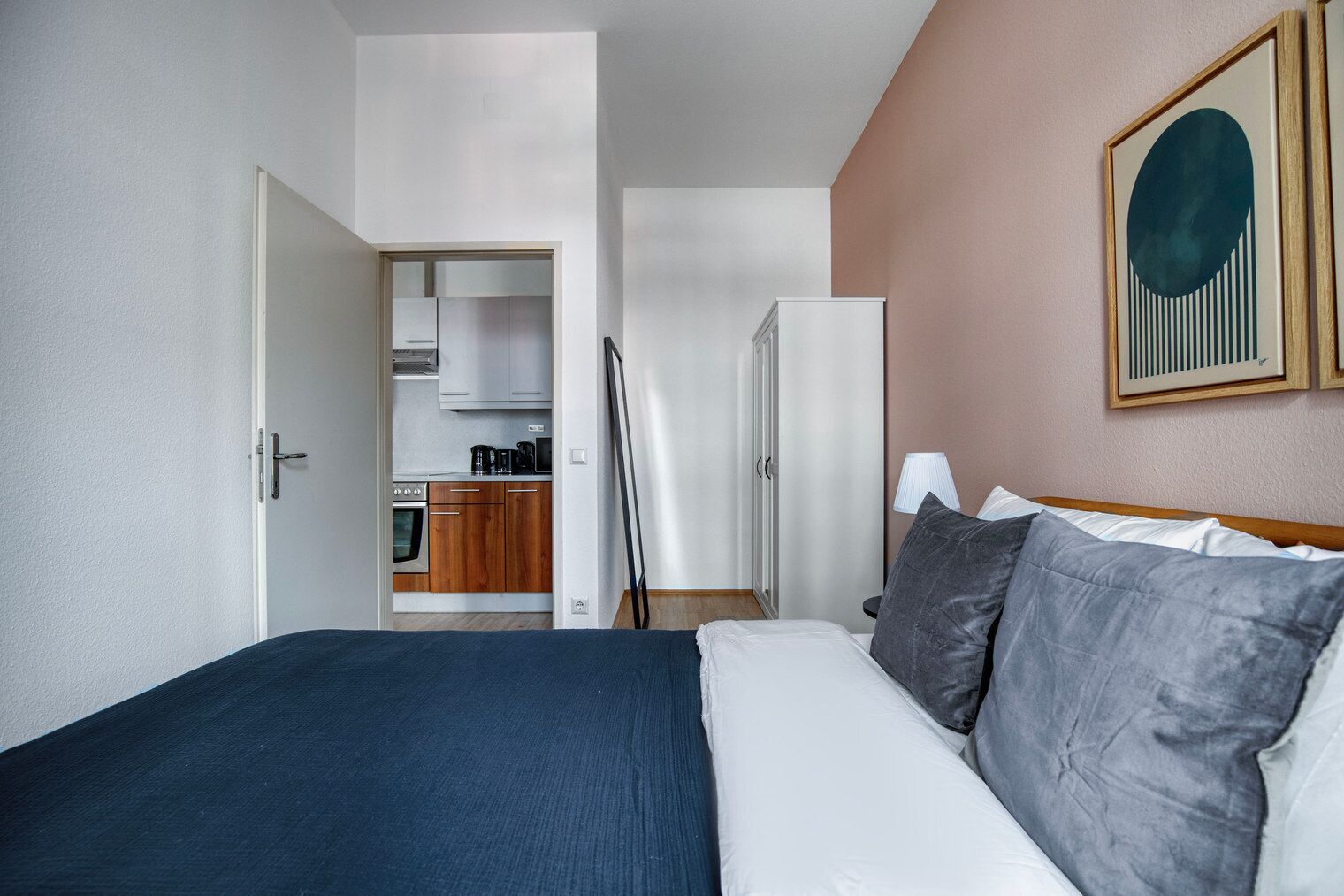 A hotel room with a bed, a desk, a chair, a wardrobe, and a kitchenette.