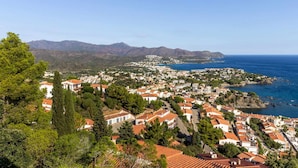 Un poble costaner amb edificis de teules vermelles, un mar blau clar i muntanyes al fons.