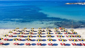 A beach with rows of beach chairs and umbrellas, a clear blue sea, and a pier in the background. - Beautiful apartment in villa for 4 guests with WIFI, pool, A/C, TV, terrace and pets allowed (Alghero)
