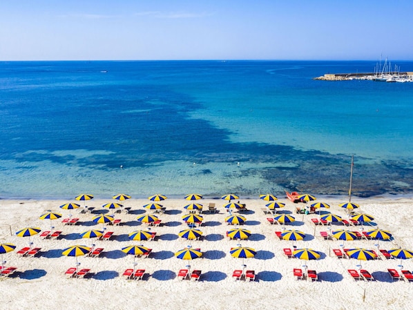 Une plage bordée de rangées de transats et de parasols, une mer d’un bleu limpide et, au loin, une jetée.