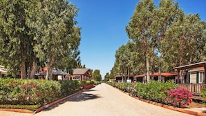 A paved pathway flanked by trees and flower beds, leading to a row of wooden cabins with red roofs. - Nice apartment in villa for 4 people with WIFI, pool, A/C, TV and terrace (Alghero)