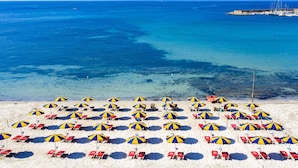 A beach with rows of beach chairs and umbrellas, a clear blue sea, and a pier in the background. - Beautiful apartment in villa for 6 guests with WIFI, A/C, pool, TV and terrace (Alghero)