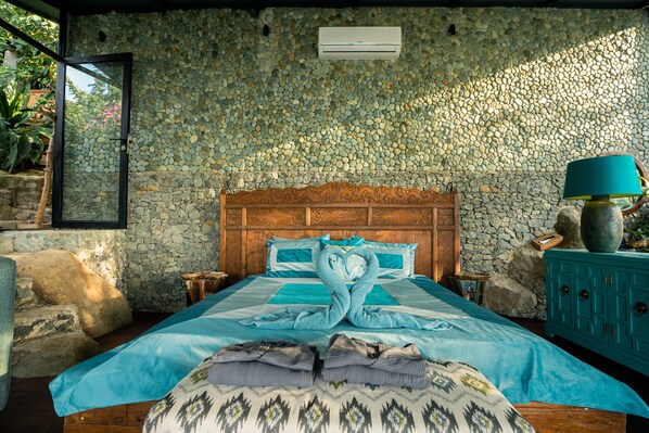 Une chambre avec un mur en pierre, une tête de lit en bois, un lit avec une literie bleue, une lampe turquoise et une table de chevet en bois.