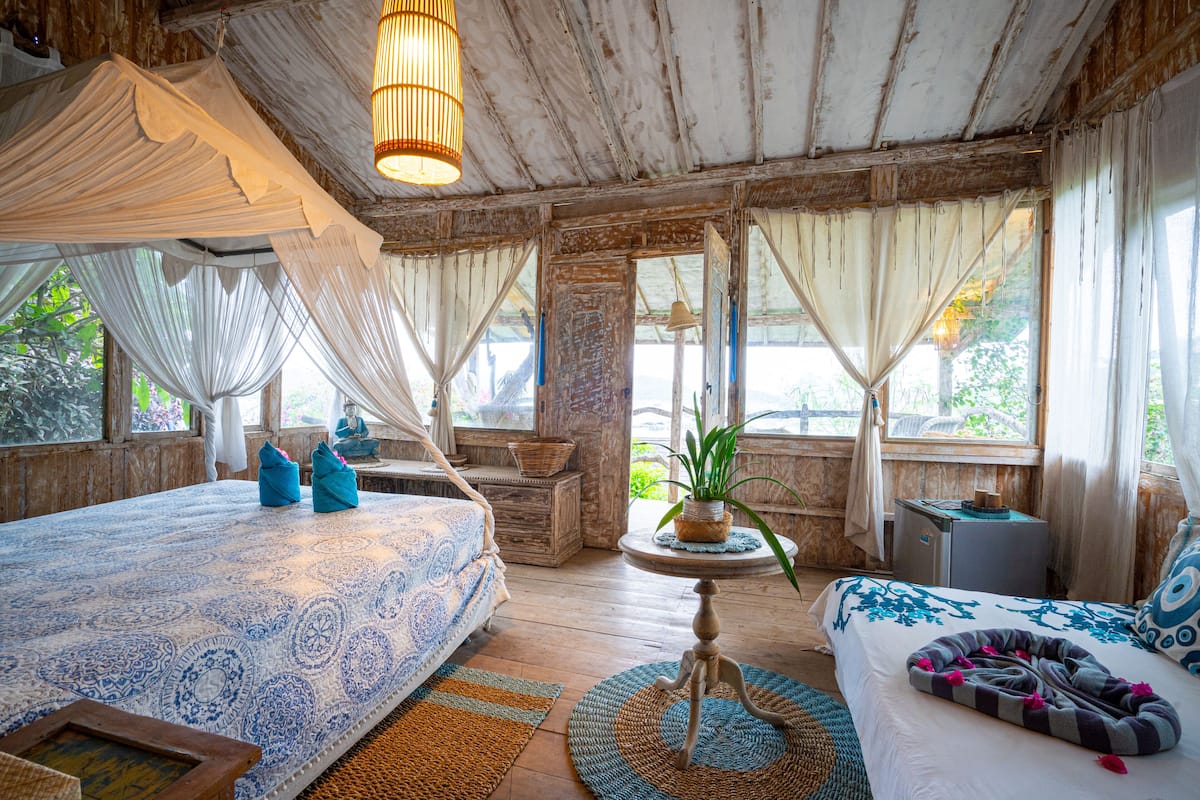 A rustic bedroom with a canopy bed, a smaller bed with a patterned bedspread, a round table with a plant, and a small nightstand.