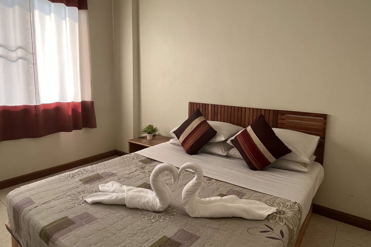 A hotel room with a bed, a nightstand, a potted plant, and a window with curtains.