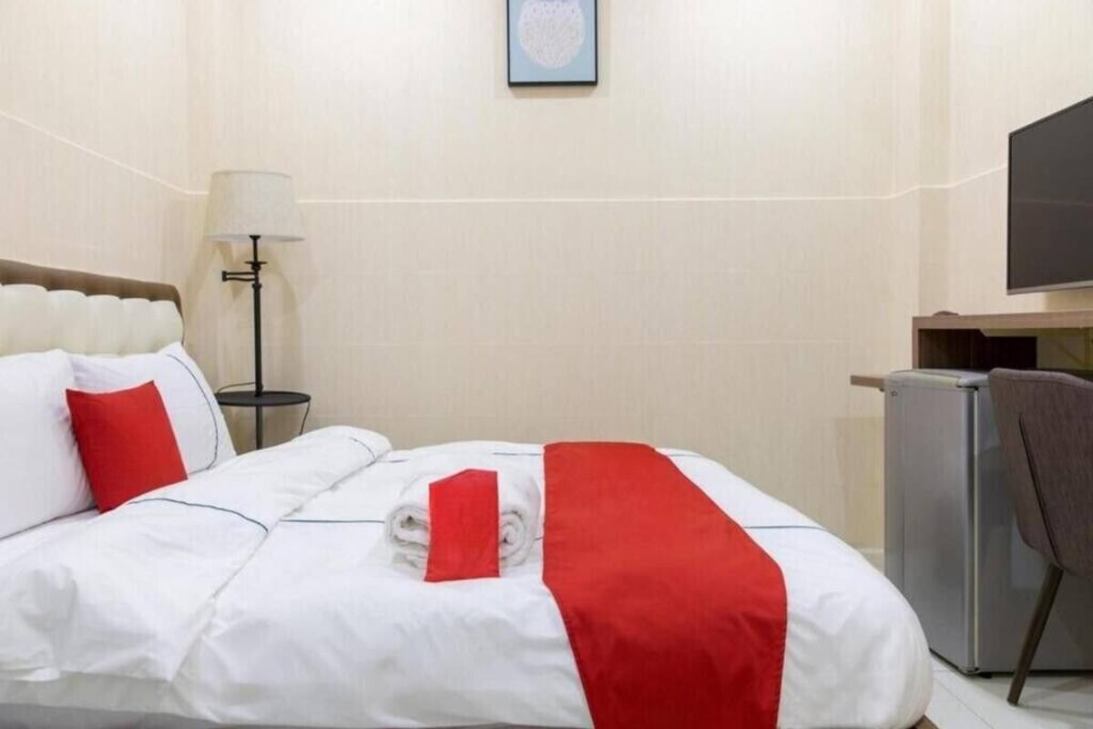 A hotel room with a neatly made bed, a desk with a flat-screen TV, a lamp, and a refrigerator.