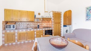 Een kleine keuken met houten kastjes, een magnetron en een spoelbak. In de aangrenzende woonkamer staat een tafel met een kom en een bank.