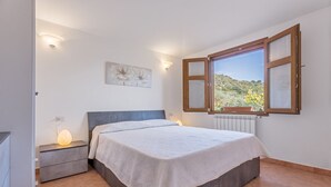 Een slaapkamer met een bed, nachtkastje, wandlamp en een raam met uitzicht op groen.