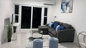 A modern living room with a grey sectional sofa, a glass coffee table, and a small side table. There is a large window with a view of the outside. - Luxury one bed room apartment with AC, roof top pool, WIFI located in Kingston. (Kingston)