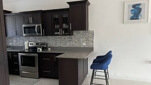 A modern kitchen with dark cabinetry, stainless steel appliances, a tiled backsplash, and a bar area with blue barstools. - Luxury one bed room apartment with AC, roof top pool, WIFI located in Kingston. (Kingston)
