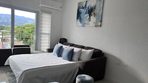 A bedroom with a bed, a sofa, two ottomans, a painting, and a view of mountains through a sliding door. - Luxury one bed room apartment with AC, roof top pool, WIFI located in Kingston. (Kingston)