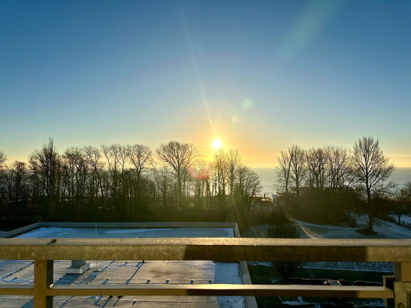 A clear view of a sunset over a body of water, with bare trees in the foreground and a building with a snow-covered roof in the background.