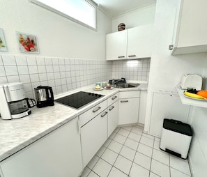 Cocina compacta con armarios blancos, cafetera, lavamanos y una pequeña exhibición de frutas.