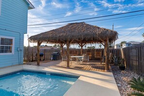 A backyard with a swimming pool, a covered patio with outdoor furniture, and a wooden fence.