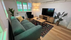 A modern living room with a green sofa, a black chair, a wooden coffee table, a flat-screen TV, and a decorative plant. - Sans Souci Apartments (Ingham)