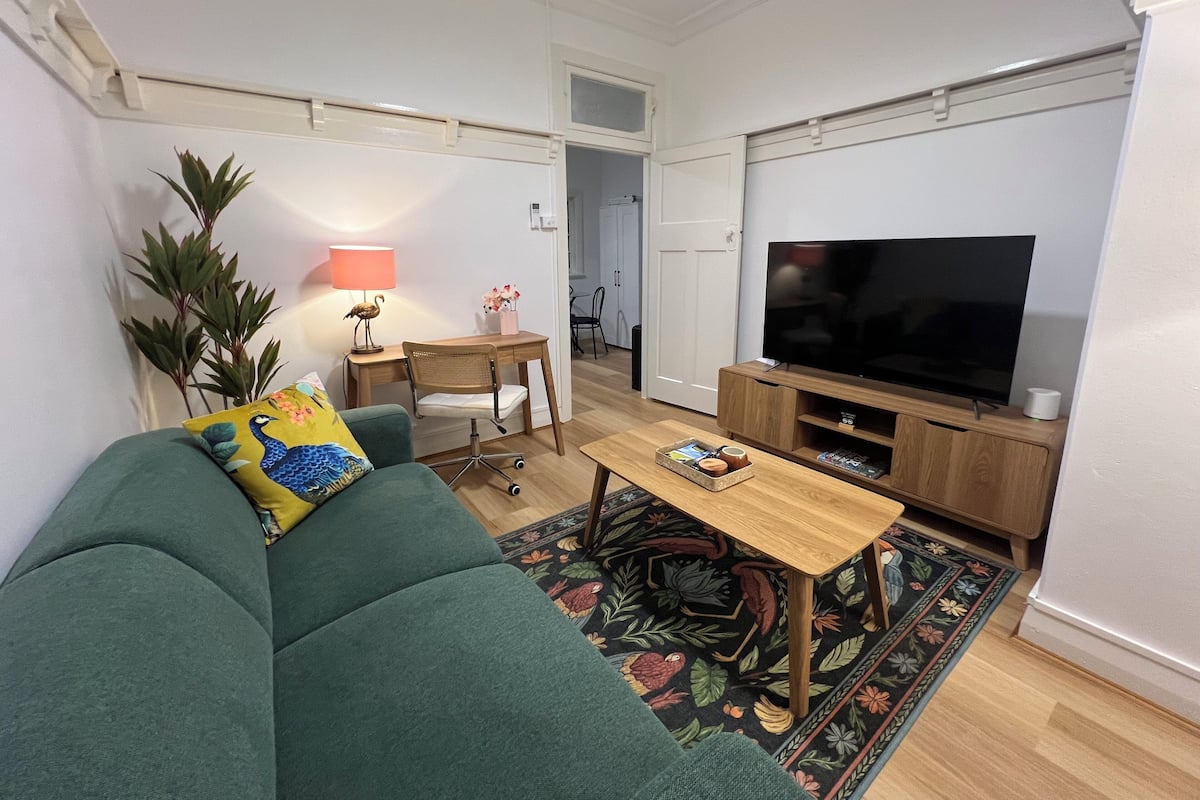 Ein Wohnzimmer mit einem grünen Sofa, einem Holztisch, einem Fernseher und einem Schreibtisch mit einer Lampe.
