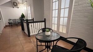 A spacious room with a tiled floor, a round glass table with a potted plant, two black chairs, a white-framed window, and a white brick wall. - Sans Souci Apartments (Ingham)
