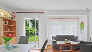 Una sala moderna con una mesa de comedor, un sofá y una mesa de centro. Hay una estantería y un ventanal con cortinas.