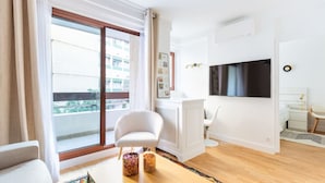 A modern living room with a large sliding glass door leading to a balcony, a flat-screen TV mounted on the wall, a white armchair, and a small table with a colorful rug. - Passy - 1 chambre - 2 lits (Paris)