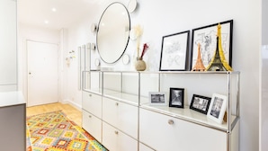 A hallway with a patterned rug, a mirror, and framed pictures on a glass cabinet. - Passy - 1 chambre - 2 lits (Paris)