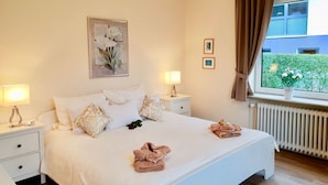 Une chambre d’hôtel avec un lit, des tables de chevet, une fenêtre avec des rideaux, un radiateur et un mur orné de tableaux.