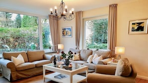 Un salon avec deux canapés, une table basse et un lustre. Il y a des fenêtres avec des rideaux, un tableau au mur et une petite table avec une lampe.