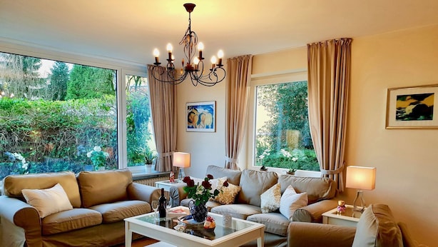 Un salon comprenant un ensemble de canapés, une table basse et un lustre. On y trouve des fenêtres avec des rideaux, un tableau au mur et une vue sur la verdure à l’extérieur.
