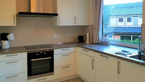 Une cuisine moderne avec des armoires blanches, un four encastré, une hotte aspirante et une fenêtre donnant sur un bâtiment au toit bleu.