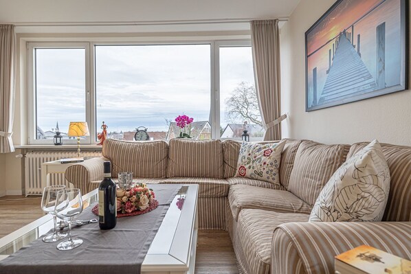 En stue med en stribet sofa, et bord med vinglas og en flaske, og et stort vindue med udsigt til bygninger.