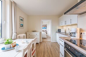 Et moderne køkken med indbygget ovn, mikroovn og kaffemaskine. Der er et spiseområde med bord og stole, og et soveværelse kan anes gennem en åben dør.