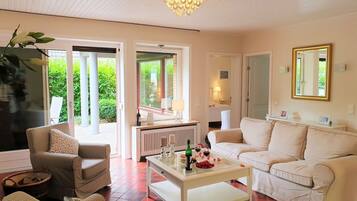 Ein geräumiges Wohnzimmer mit einem weißen Sofa, einem Glastisch und einem Kronleuchter. Eine Tür führt zu einem Balkon mit Blick auf Grünflächen.