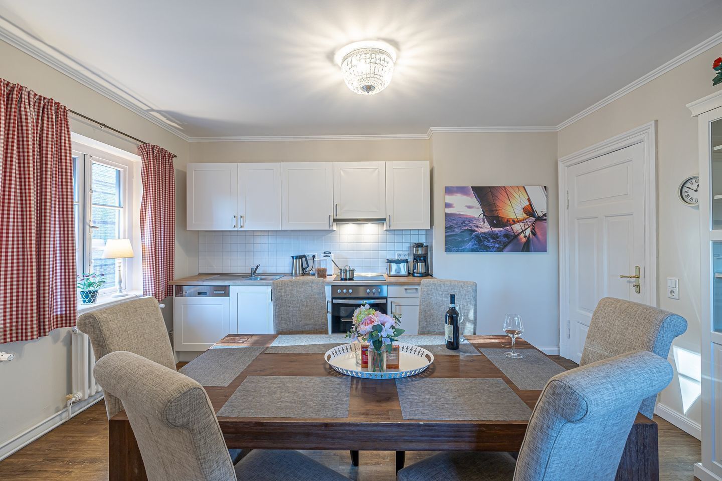 Una cocina moderna con armarios blancos, un comedor con mesa de madera y un cuadro de un velero en la pared.